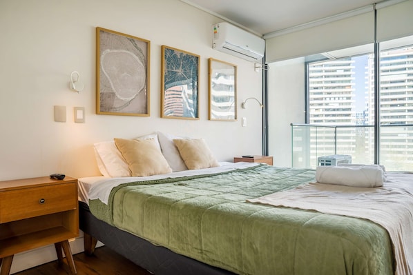 A modern bedroom with a large bed, a nightstand, a bedside table, and a window with a view of buildings.