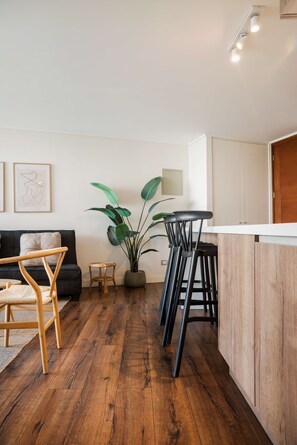 A modern living space with a black sofa, wooden floor, a dining area with stools, a potted plant, and a white wall with framed artwork.