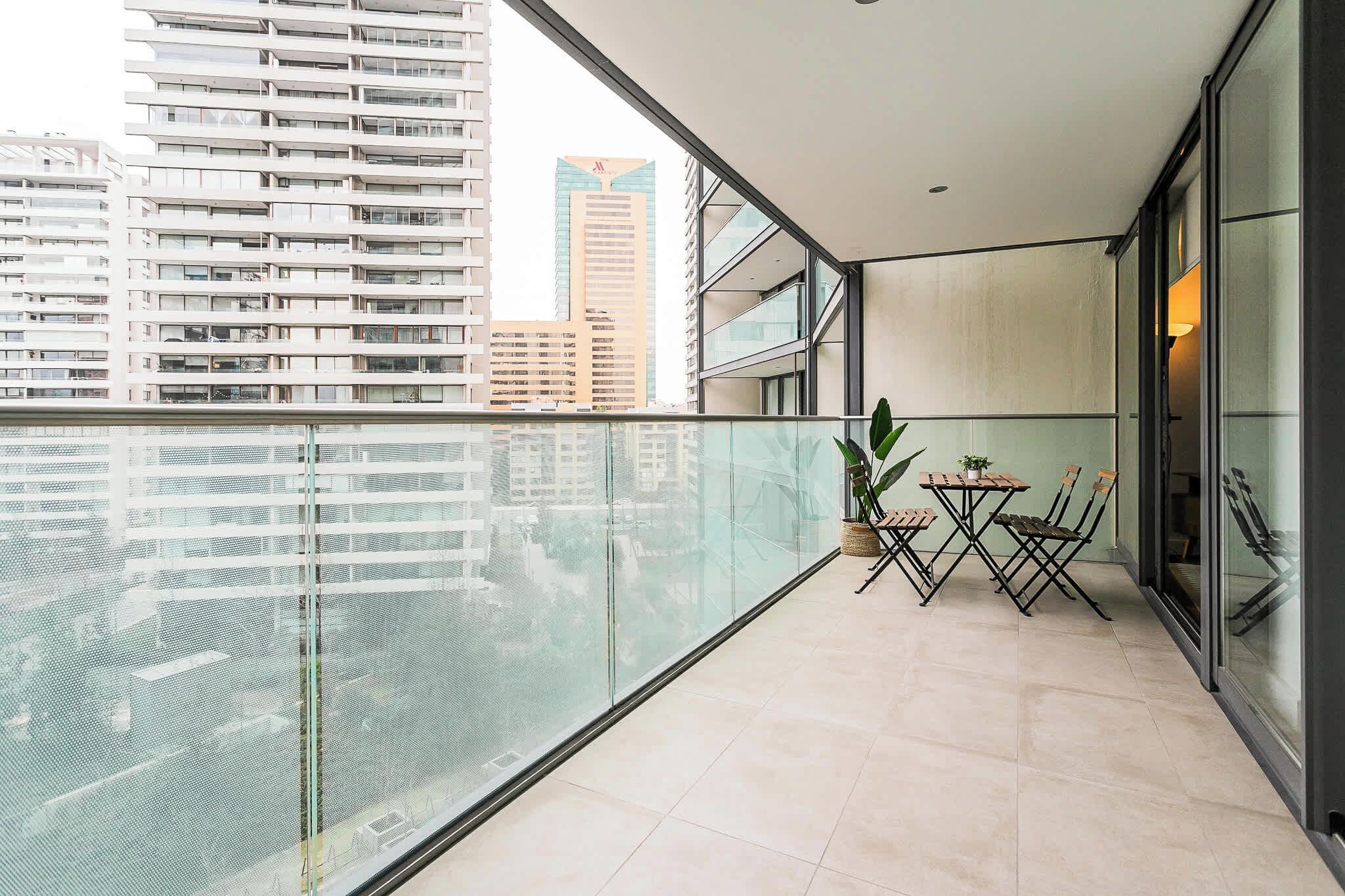 A balcony with a glass railing, a small table with chairs, a potted plant, and a view of tall buildings.