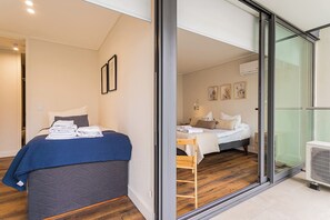 Ein modernes Schlafzimmer mit einer großen Glaswand, die auf einen Balkon führt, einem Bett mit blauer Bettdecke und einem weiteren Bett mit weißer Bettdecke.