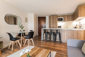 A modern kitchen with wooden cabinets, a central island with bar stools, a dining area with a glass table and chairs, and a small seating area with a sofa.