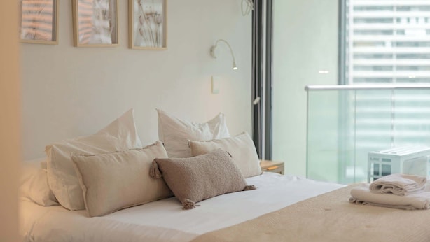 A hotel room with a bed, pillows, a bedside table, a lamp, and a view of the city through large windows. - Chic Las Condes Apt: 2BR, 2BA, Sleeps 5 (Las Condes)