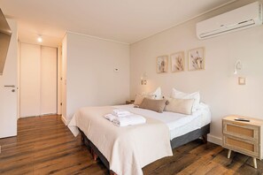Une chambre à coucher comprenant un lit, une table de chevet, un climatiseur fixé au mur et une porte donnant sur une autre pièce.