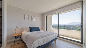 Une chambre à coucher avec un lit, une table de chevet, une lampe et une vue sur le paysage urbain, visible à travers de grandes fenêtres.