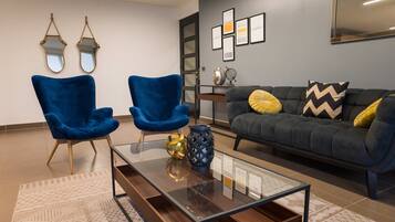 A modern living room with two blue armchairs, a dark grey sofa with yellow and black chevron pillows, a glass coffee table with decorative vases, and a wall with framed artwork and mirrors.