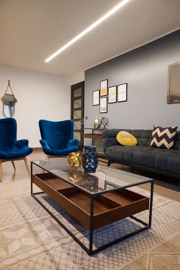 Ein modernes Wohnzimmer mit einem Glastisch, zwei blauen Sessel, einem grauen Sofa mit dekorativen Kissen und gerahmten Kunstwerken an der Wand.
