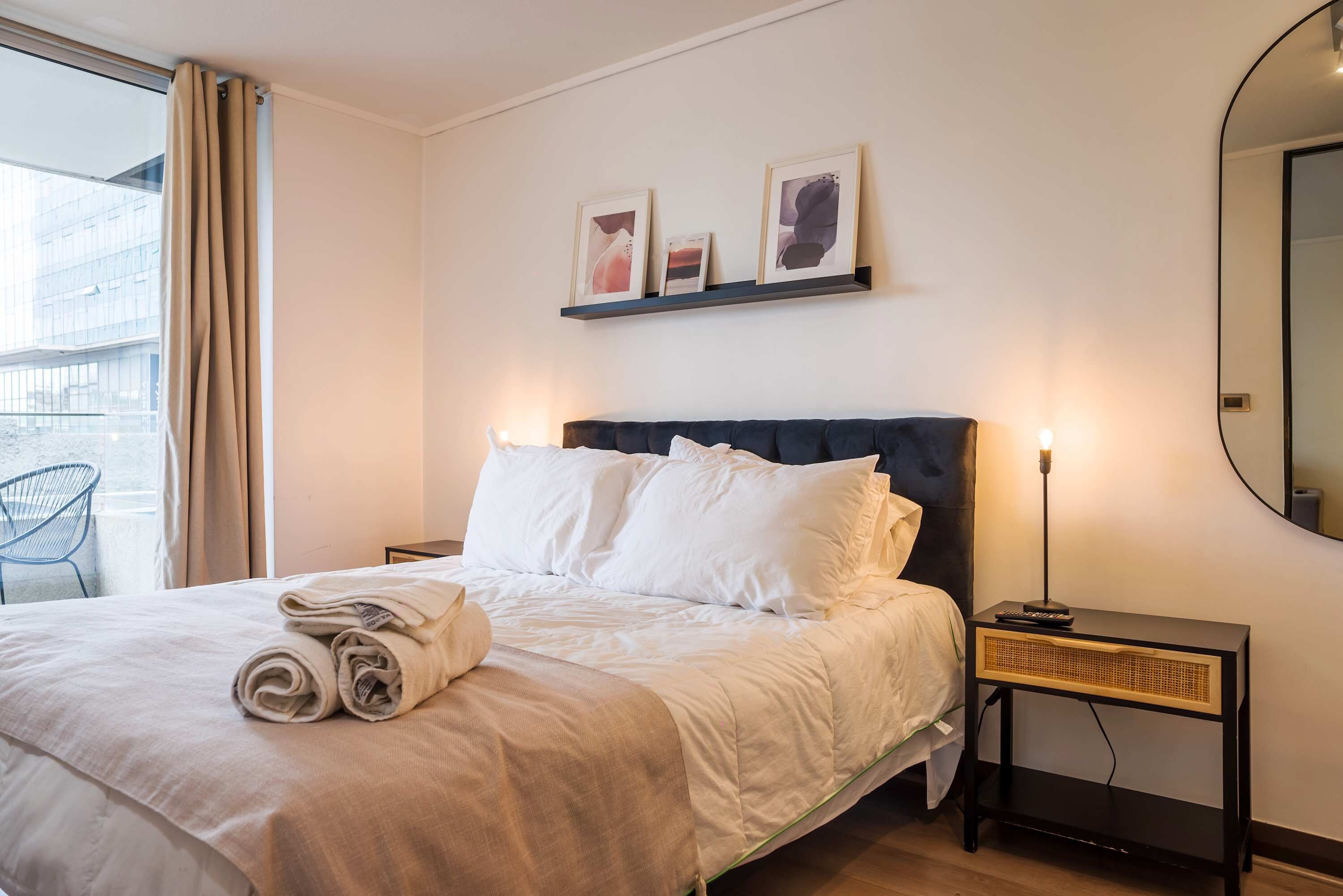 A hotel room with a bed, a nightstand, a lamp, and a wall-mounted shelf with pictures.