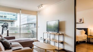 Una sala moderna con un sofá, una mesa de centro, un televisor sobre un mueble bajo y una cama con una mesita de noche.