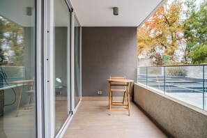 A balcony with a wooden chair and a table, a glass door leading to an interior space, and a view of trees with autumn foliage.