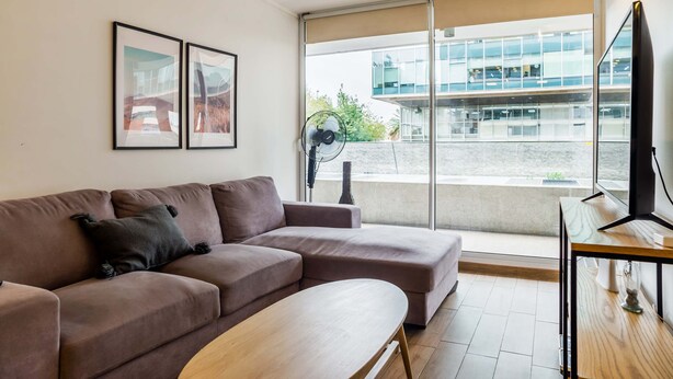 Una sala de estar moderna con un sofá, una mesa de centro y un televisor. Hay dos cuadros enmarcados en la pared y un ventanal con vistas a un edificio.