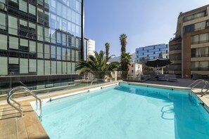 Une piscine sur le toit, entourée d’une barrière en verre, avec des palmiers et des bâtiments modernes en arrière-plan.