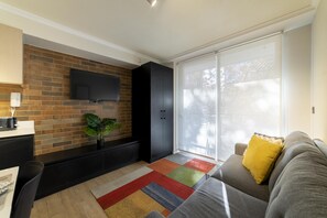 Un salon moderne avec un téléviseur à écran plat fixé à un mur en briques, un meuble noir, un tapis coloré et un canapé gris agrémenté d’un coussin jaune.