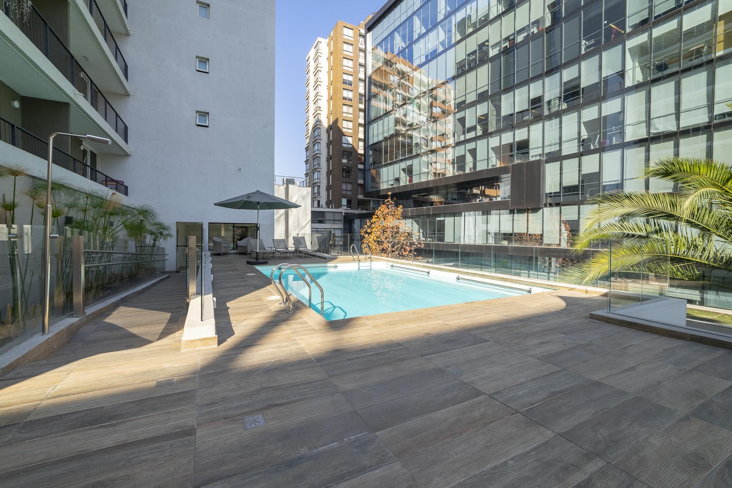 A rooftop pool area with a glass balustrade, a water feature, and a view of surrounding high-rise buildings.