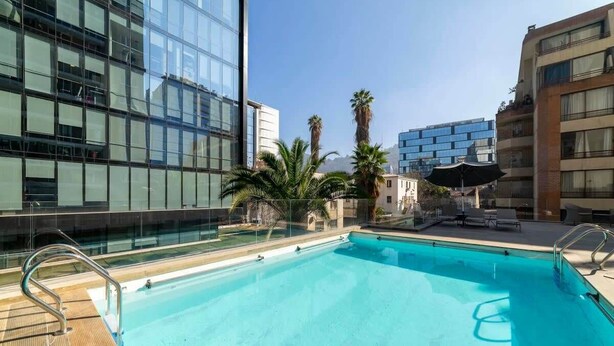 Una piscina en la azotea con agua azul cristalina, rodeada de edificios modernos y palmeras.