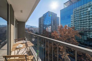 A balcony with a wooden table and chairs, overlooking a cityscape with modern buildings and autumn trees.