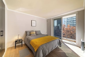 Private bedroom with a mustard throw, minimalist styling, and floor-to-ceiling glass doors opening to a balcony with autumn trees and mountain views.