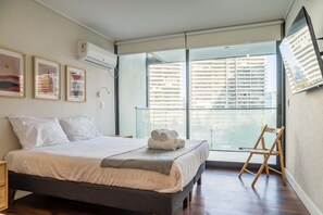Une chambre moderne avec un grand lit, un fauteuil et une vue sur un immeuble de grande hauteur, visible à travers de grandes fenêtres du sol au plafond.
