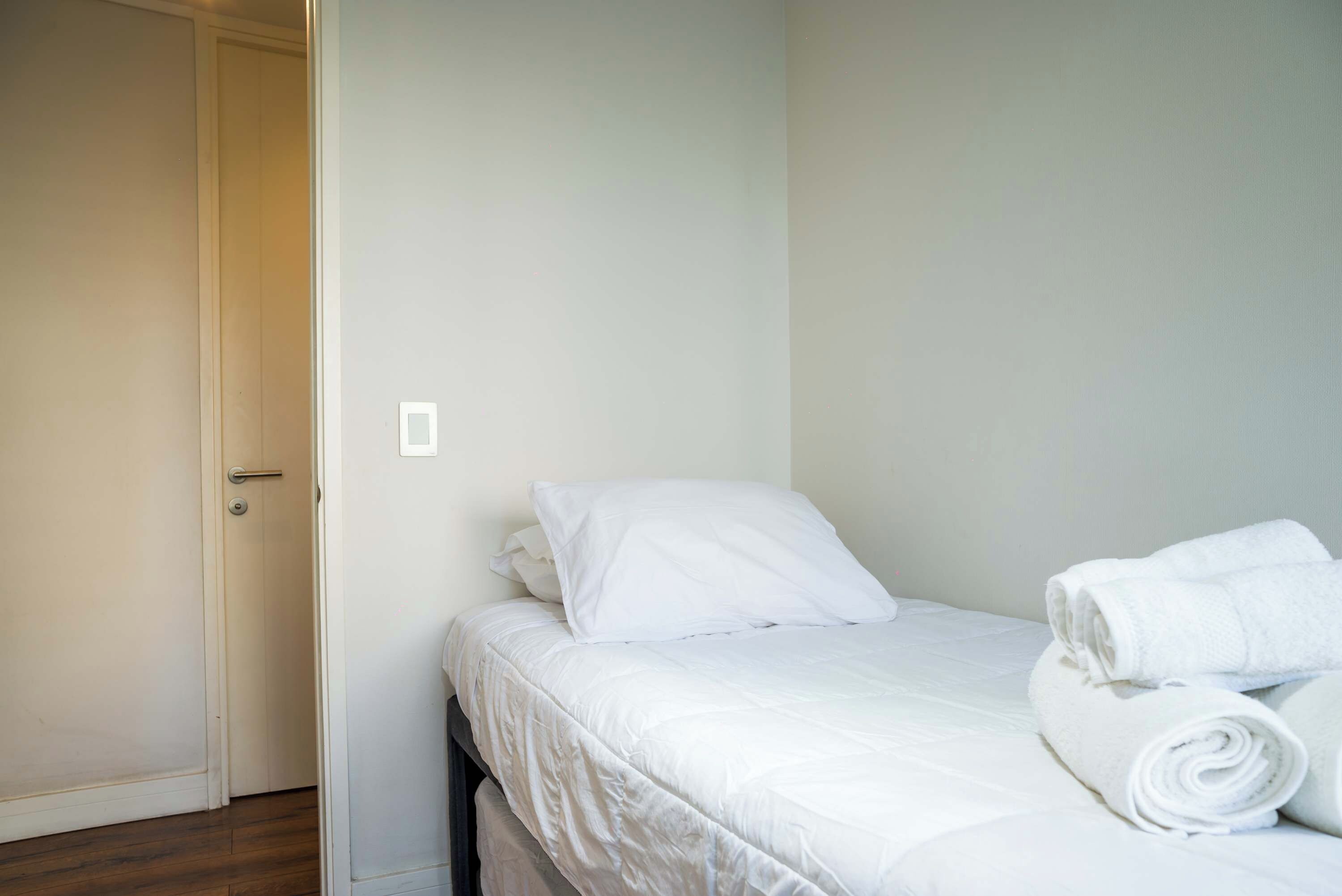 Une chambre avec un lit simple, une porte, un interrupteur et une serviette enroulée posée sur le lit.