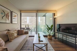 A modern living room with a sofa, coffee table, TV, and a balcony with a view of high-rise buildings. - Comfortable Apartment in Las Condes: Pool and Gym! (Las Condes)