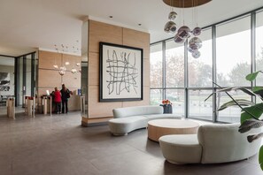 Un hall d’entrée moderne avec une grande peinture abstraite, un canapé blanc aux lignes courbes et une table basse ronde. Des portes vitrées donnent accès à un autre espace, et une grande fenêtre offre une vue sur l’extérieur.