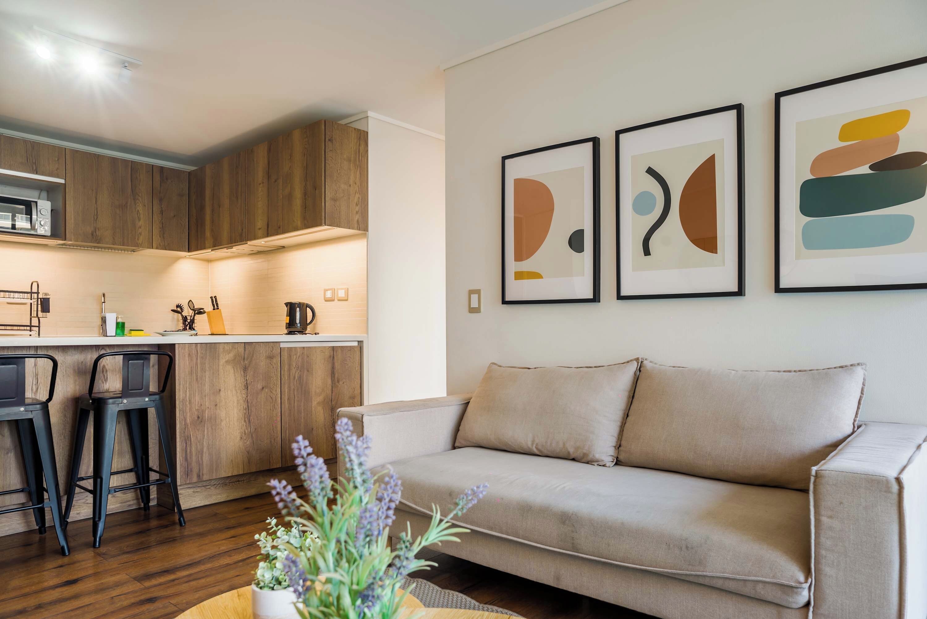 Een moderne woonkamer met een bank, twee zwarte barkrukken, een salontafel met een plant, en een keuken met houten kastjes en een magnetron.