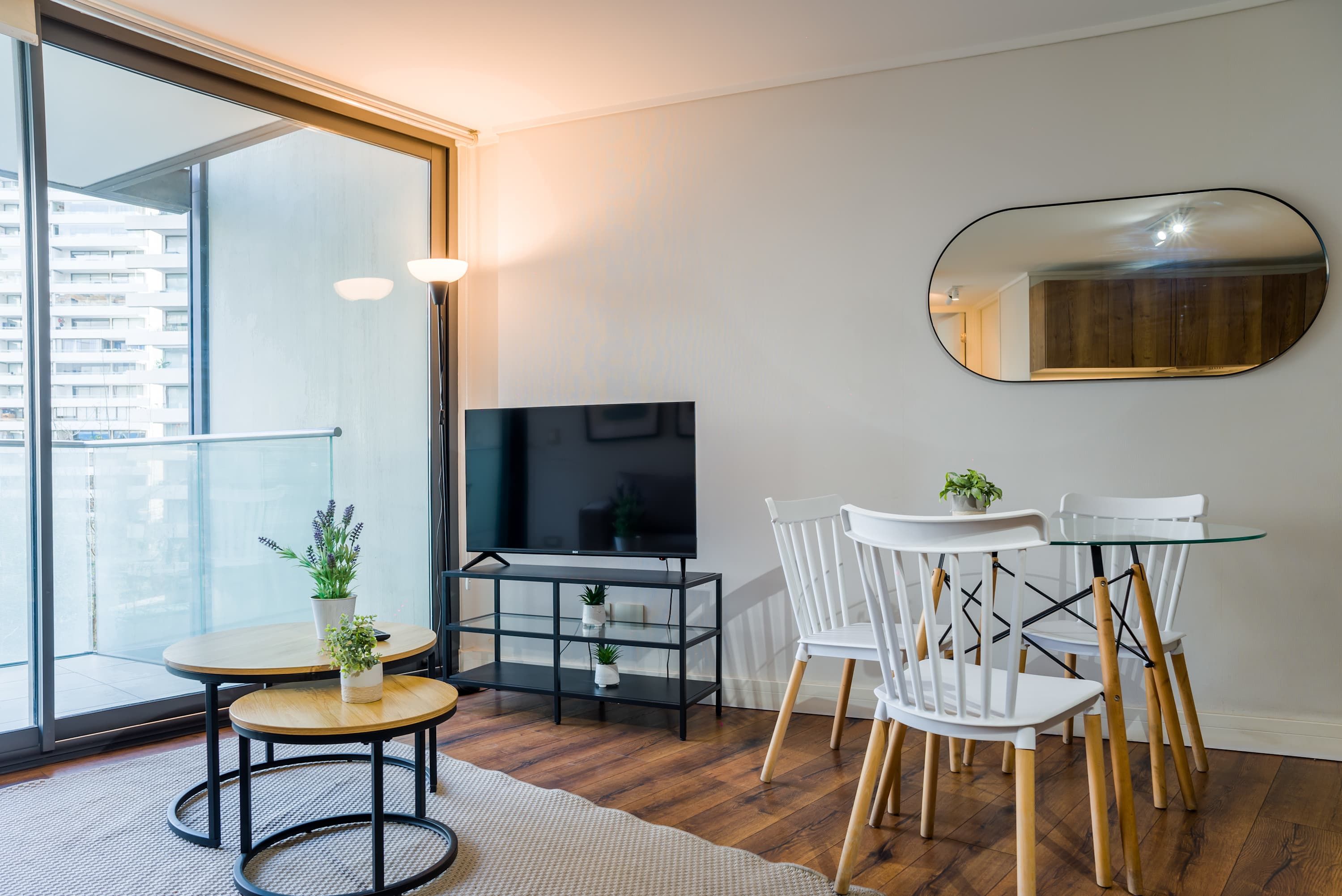 Ein modernes Wohnzimmer mit Fernseher, einem Glastisch, weißen Stühlen und einem großen Fenster.