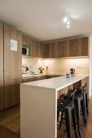 A modern kitchen with wooden cabinets, a central island with bar stools, a microwave, and a coffee maker. - Comfortable Apartment in Las Condes: Pool and Gym! (Las Condes)