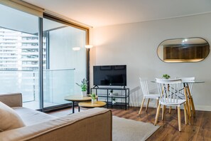 A modern living room with a sofa, a small round table with a plant, a TV on a stand, and a dining area with chairs and a table. - Comfortable Apartment in Las Condes: Pool and Gym! (Las Condes)