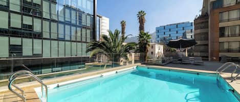 A rooftop pool with clear blue water, surrounded by modern buildings and palm trees.