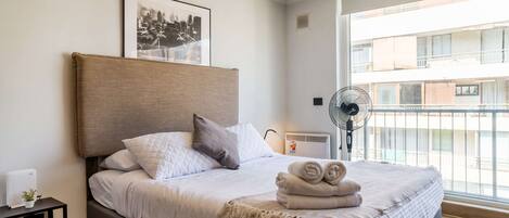 A neatly made bed with a headboard, pillows, a blanket, and rolled towels. There is a framed picture on the wall, a small table with a plant, and a standing fan.