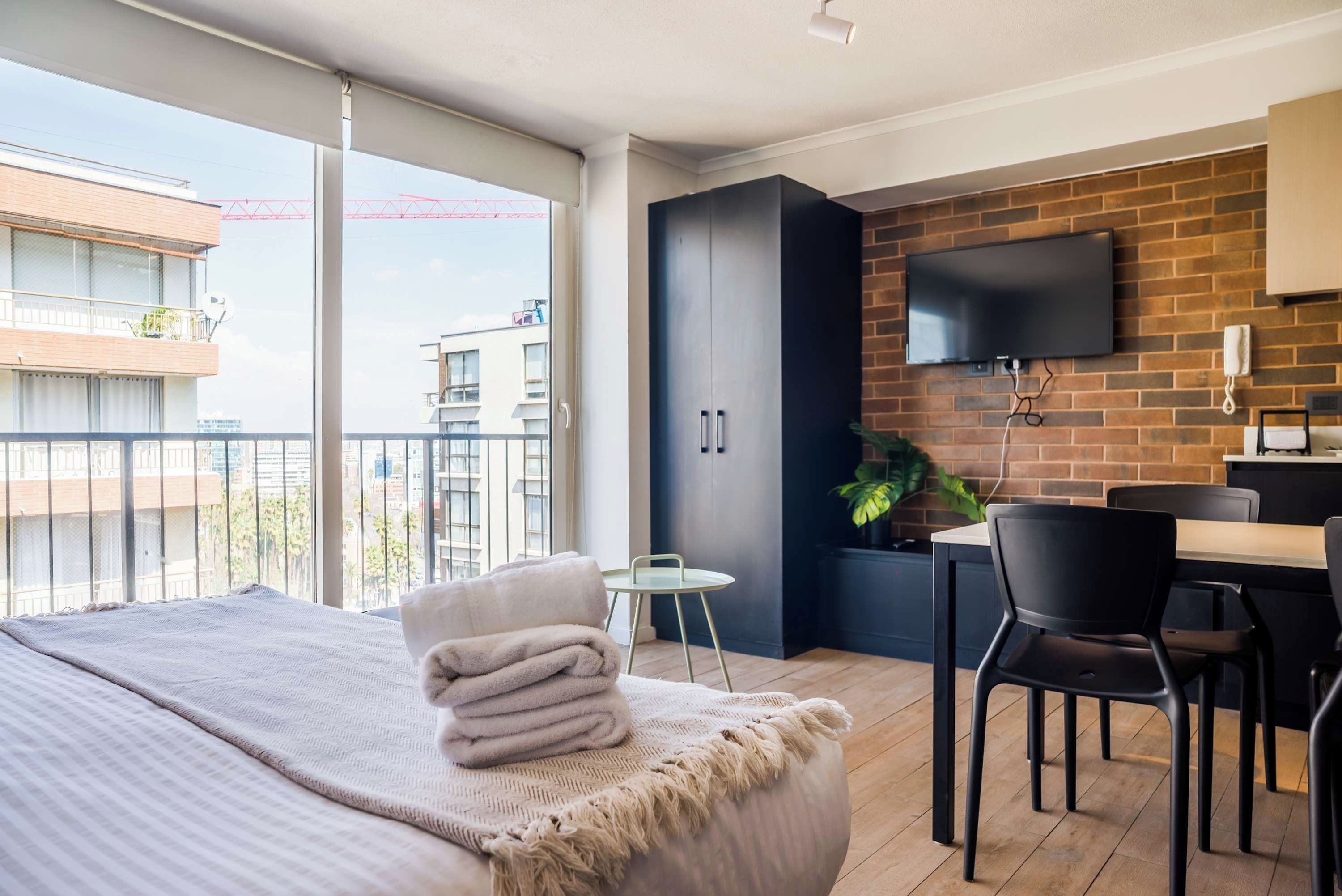 Ett modernt hotellrum med en stor säng, ett skrivbord, en stol, en tv som är monterad på en tegelvägg och en balkong med utsikt över byggnaderna.