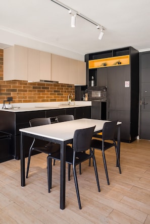 A modern kitchen with a brick backsplash, a dining area with a white table and black chairs, and a black cabinet with a yellow shelf.