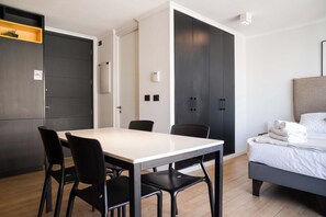 A modern hotel room with a bed, a dining table with chairs, a kitchenette, and a wardrobe.