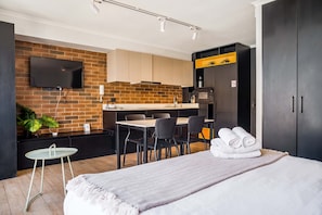 A modern hotel room with a bed, a small table, a desk, and a kitchen area with a brick wall and a flat-screen TV. - Stylish and Cozy Studio Apartment in Providencia (Providencia)