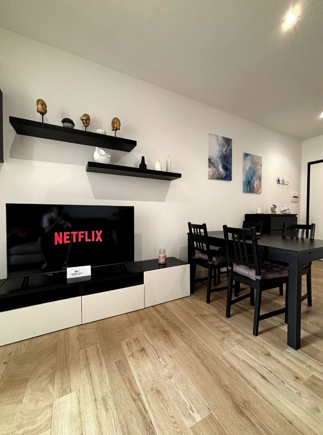 Un salon moderne doté d’un téléviseur à écran plat, de planchers en bois, d’un coin repas avec une table et des chaises aux couleurs sombres, et d’objets décoratifs disposés sur des étagères.
