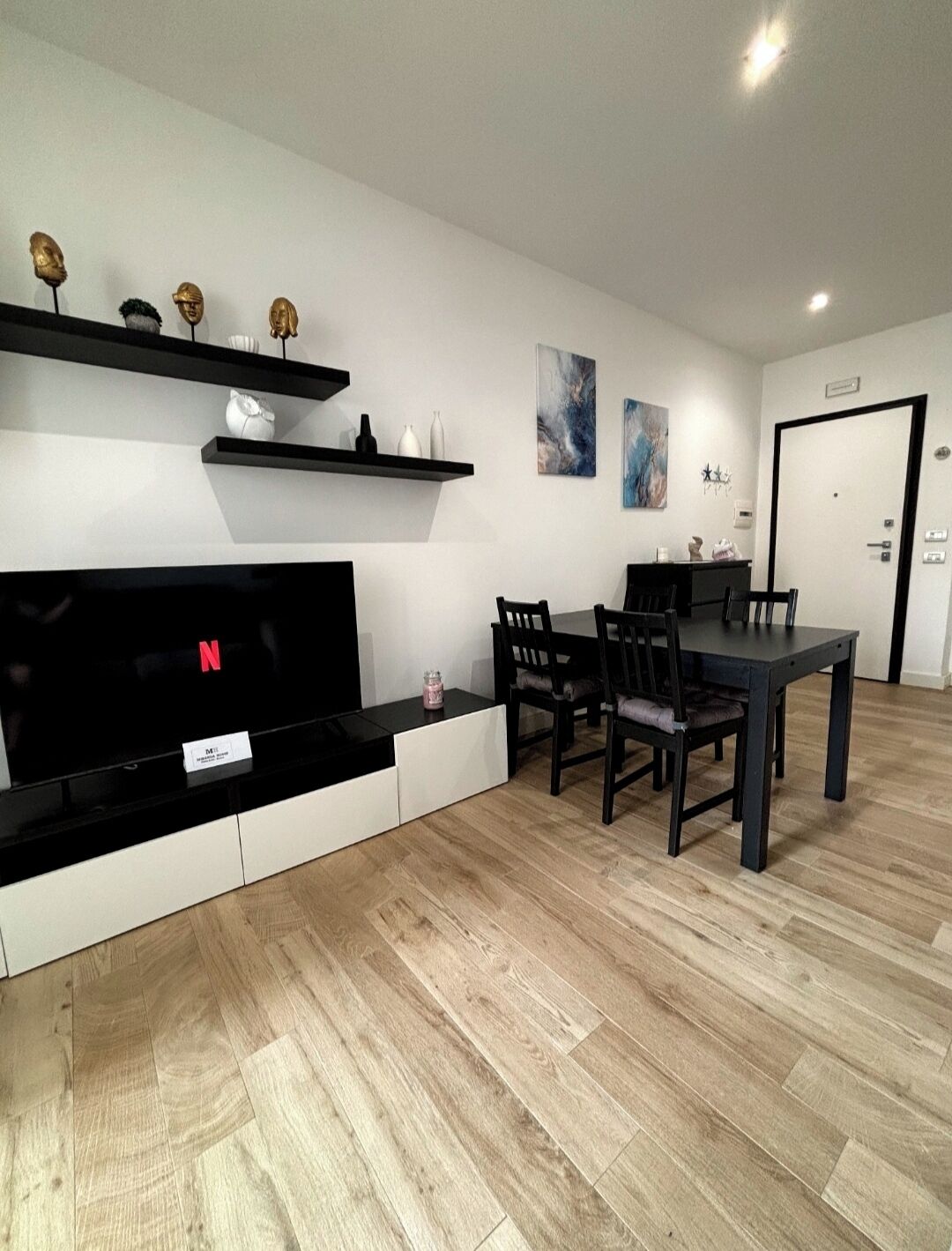 A modern living room with a flat-screen TV, black and white entertainment unit, wooden flooring, and a dining area with a dark table and chairs.