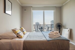 Een slaapkamer met een bed, kussens, een ventilator, een radiator en een balkon met uitzicht op gebouwen.