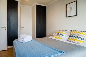 Una habitación de hotel con una cama, una manta azul, dos almohadas con diseños en zigzag, un cuadro en la pared y una puerta.