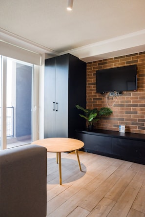 Un intérieur moderne avec une table à manger en bois, un meuble noir, un téléviseur fixé sur un mur en briques et une porte-fenêtre coulissante donnant sur un espace extérieur.