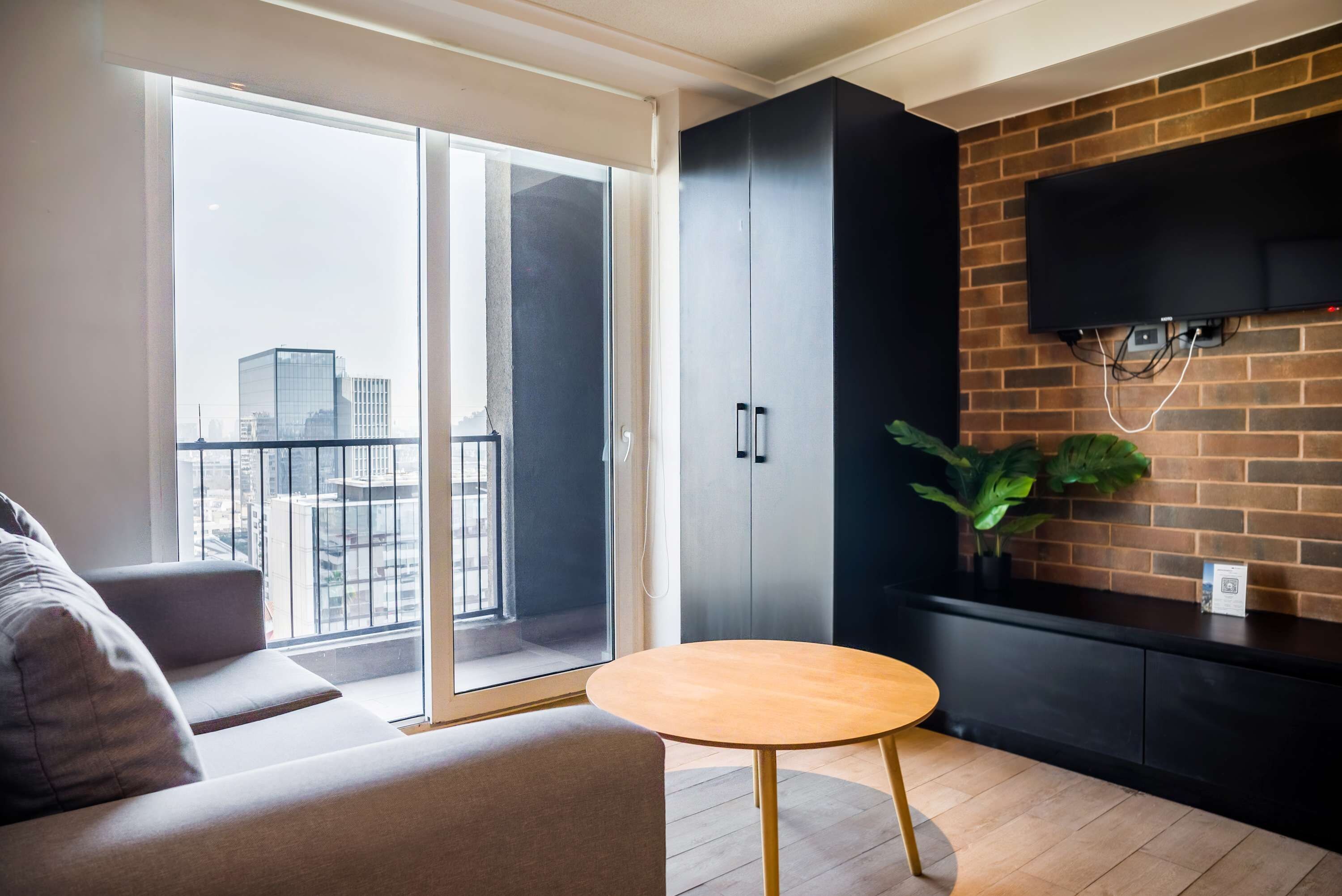 A modern living room with a sofa, a round wooden table, a sliding glass door leading to a balcony, a black cabinet, and a mounted TV.
