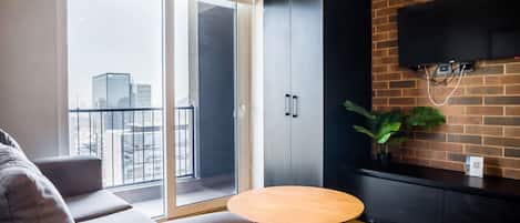 A modern living room with a sofa, a round wooden table, a sliding glass door leading to a balcony, a black cabinet, and a mounted TV.