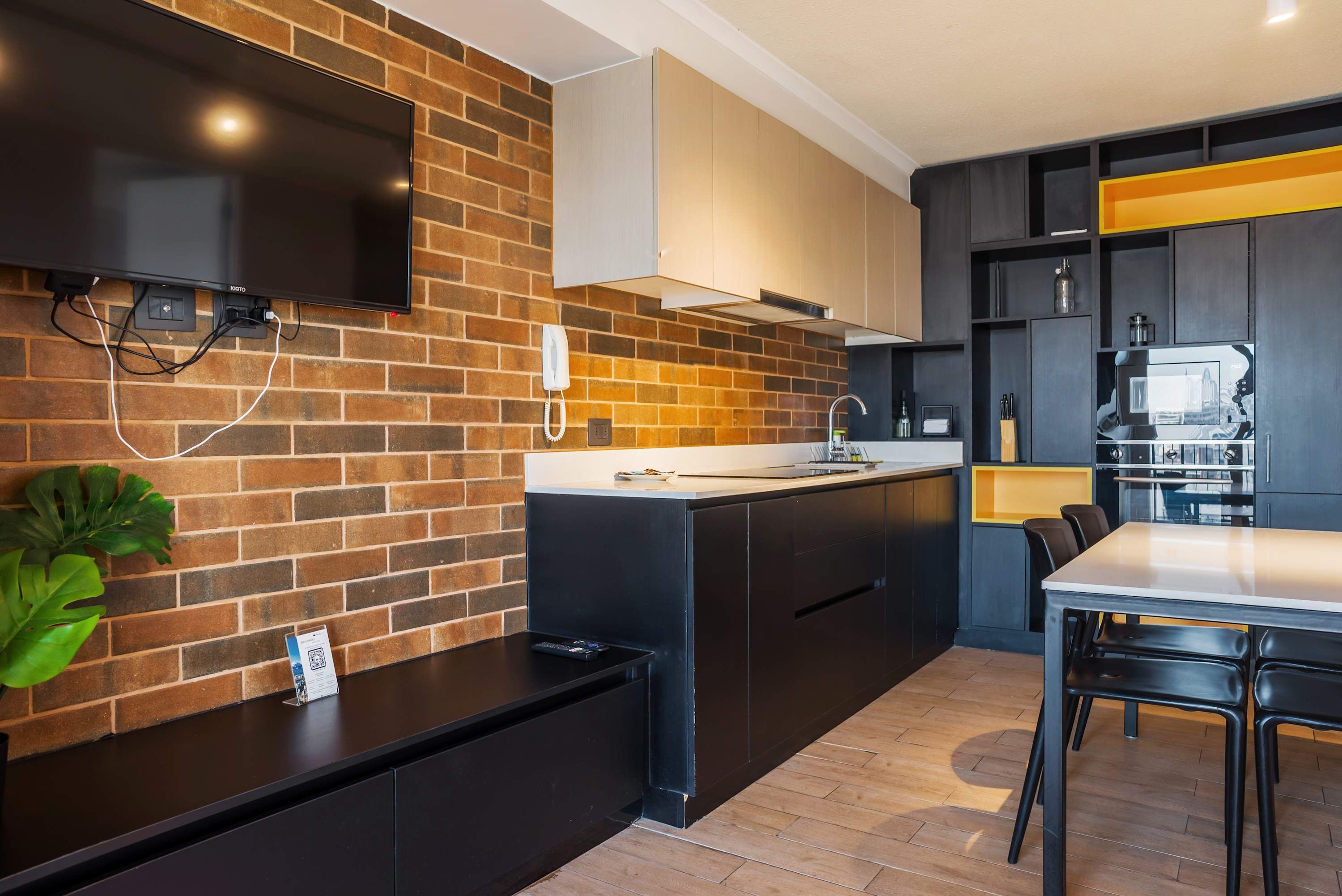 A modern kitchen with a brick wall, a flat-screen TV mounted above the counter, a built-in phone, and a dining area with a table and chairs.