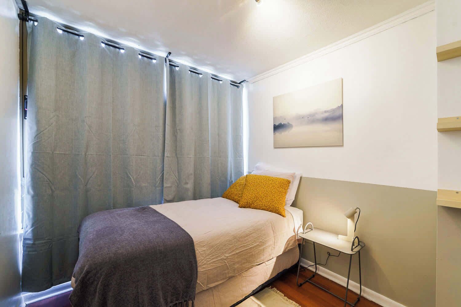 Ein Schlafzimmer mit einem Bett, einem Nachttisch, einem Wandbild und einem Fenster mit Vorhängen.