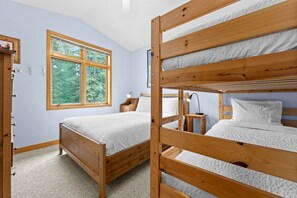 A room with two wooden bunk beds, a window with a view of trees, a bedside table with a lamp, and a wooden wardrobe.