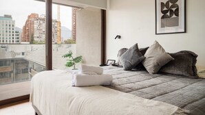 A modern bedroom with a large bed, pillows, a framed abstract art piece, and a view of the city through a balcony. - Chic Las Condes Apt: 2BR, 2BA, Pool & Gym (Santiago)