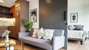 A modern living room with a grey sofa, a glass coffee table, and a plant. There is a kitchen area with wooden cabinets and a sink. The room has a grey accent wall and artwork on the wall. - Chic Las Condes Apt: 2BR, 2BA, Pool & Gym (Santiago)