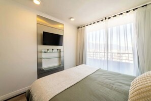 A bedroom with a bed, a mounted TV, and a window with sheer curtains.