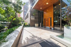 A modern building entrance with a glass door, a brick wall, and a small outdoor area with a bench and greenery.