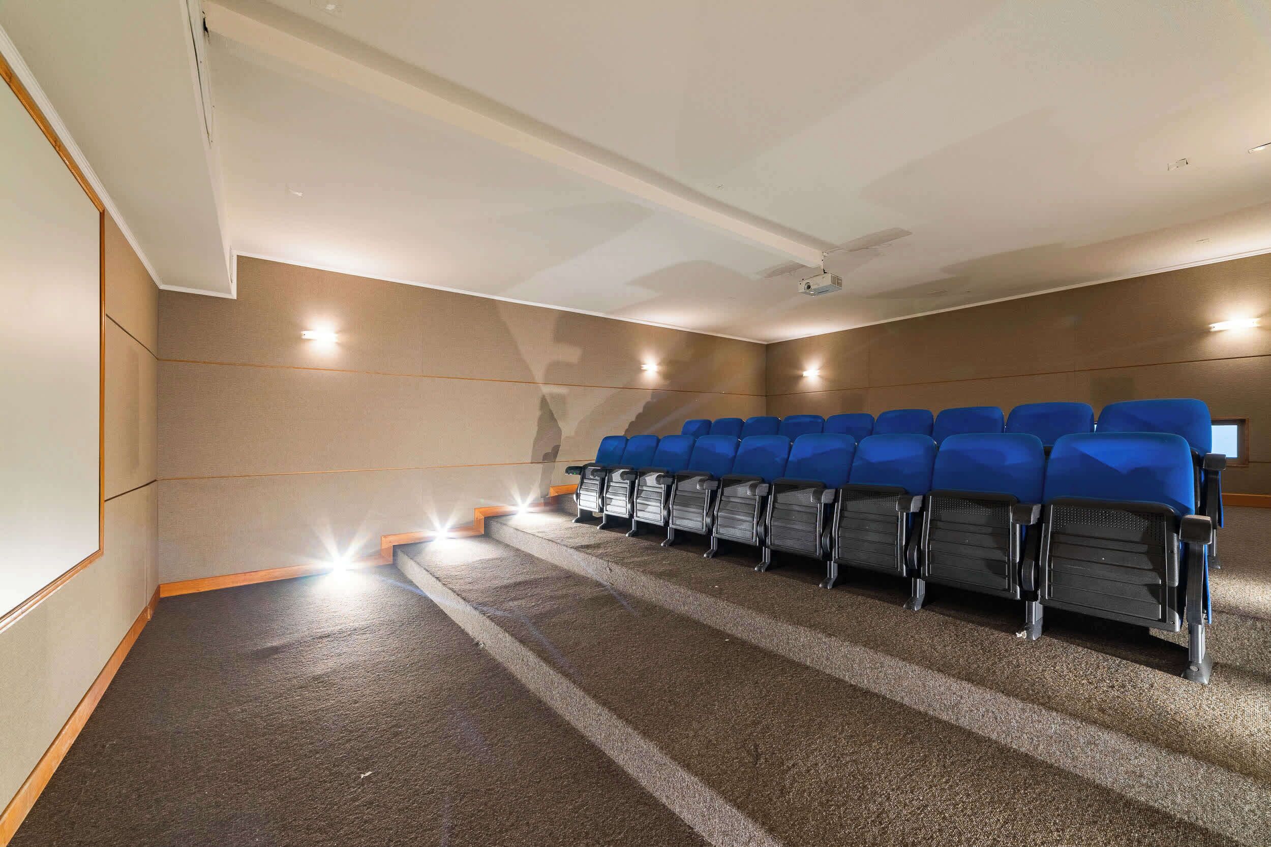 Una sala de cine con hileras de asientos azules, piso alfombrado y una pantalla de proyector.