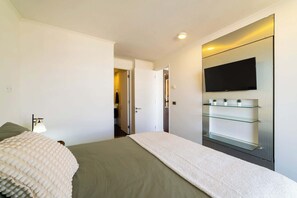 Une chambre à coucher moderne avec un lit, un téléviseur fixé au mur et une étagère en verre ornée de plantes.